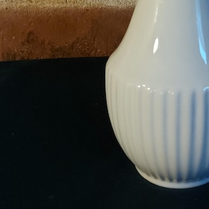 Könnte beinhalten: Eine hohe, weiße Keramikvase mit schmalem Hals und bauchiger Basis. Die Basis hat vertikale Rippendetails. Die Vase steht vor einem Hintergrund aus Backsteinmauerwerk.