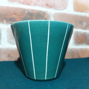 Può includere: Un vaso di ceramica color verde acqua con strisce verticali bianche. Il vaso ha una forma leggermente svasata e una superficie strutturata. Lo sfondo presenta un muro di mattoni e una superficie verde acqua scuro.