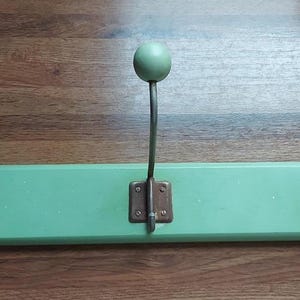 Könnte beinhalten: Ein mintgrüner Kleiderhaken aus Holz mit drei Metallhaken. Jeder Haken hat einen gebogenen, silberfarbenen Arm und einen runden, mintgrünen Knauf. Die Haken sind mit bronzefarbenen Metallplatten an der Holzbasis befestigt.