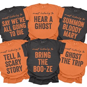 May include: Six short-sleeved crew neck t-shirts in orange and charcoal gray. Each shirt features the text "most likely to" in cursive above block-letter phrases: "SAY WE'RE ALL GOING TO DIE", "HEAR A GHOST", "SUMMON BLOODY MARY", "TELL A SCARY STORY", "BRING THE BOO-ZE", and "GHOST THE TRIP".