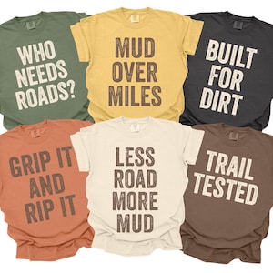 May include: Six short-sleeved t-shirts in various colours, each with a different slogan in bold, distressed lettering. Colours include olive green, mustard yellow, charcoal, rust, cream, and brown. The shirts feature phrases like "Who Needs Roads?" and "Built for Dirt."