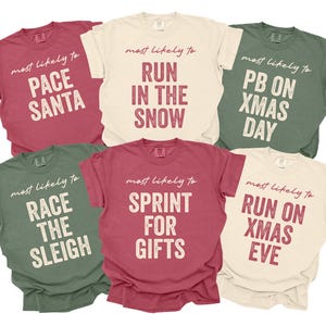 May include: A collection of six short-sleeved t-shirts in various colors, each with a different Christmas-themed phrase printed in large, bold letters. The phrases include "Pace Santa," "Run in the Snow," and "PB on Xmas Day."