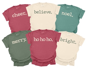 May include: Six short-sleeved t-shirts in various colors, each with a different holiday-themed word printed in a contrasting color. The words include "cheer," "believe," "noel," "merry," "ho ho ho," and "bright."