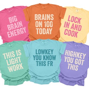 May include: Six brightly coloured t-shirts with white text. The shirts are in pink, orange, yellow, green, blue, and purple. Phrases include "Big Brain Energy," "Brains on 100 Today," and "Lock in and Cook."