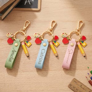 Può includere: Tre portachiavi personalizzati con hardware color oro. Ogni portachiavi presenta un cinturino in pelle colorata con un nome, un ciondolo a forma di matita, una mela rossa e un ciondolo a forma di tavolozza. I cinturini sono verdi, blu e rosa.