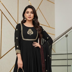 Peut inclure: Longue robe noire à manches longues avec broderies dorées et une écharpe assortie. La robe présente une jupe plissée et un motif doré décoratif sur le corsage. Une pochette dorée est tenue à la main. Le texte "kridha FASHION" est visible.