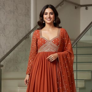 Puede incluir: Un vestido formal de color óxido con mangas largas y una falda fluida. El corpiño presenta intrincados bordados dorados alrededor del escote y las mangas. Una bufanda a juego cae sobre el hombro. El texto "kridha FASHION" es visible.