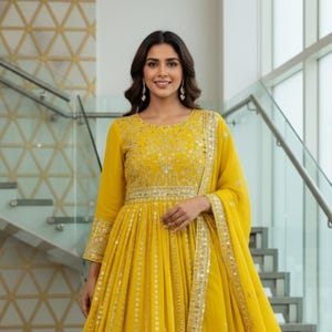 May include: A woman wearing a long, yellow dress with intricate embroidery and embellishments. The dress features long sleeves and a flowing skirt, with a matching scarf. The text "kridha FASHION" is visible on the bottom left.