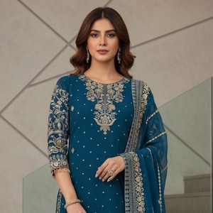 Puede incluir: Salwar kameez bordado de color turquesa con detalles dorados. El conjunto incluye un dupatta a juego y un clutch dorado. El texto "kridha FASHION" es visible en la parte inferior de la imagen.