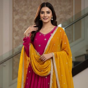 May include: A magenta-colored dress with long sleeves and a V-neck, adorned with silver star-shaped embellishments. A golden-yellow scarf with silver trim is draped over the shoulders. The dress features the text "kridha FASHION" at the bottom.