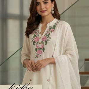 K&ouml;nnte beinhalten: Cremefarbener bestickter Kurta-Anzug mit passendem Dupatta. Der Kurta hat ein florales Design in Rosa und Gr&uuml;n um den Halsausschnitt. Der Text "kridha FASHION" ist auf dem Kleidungsst&uuml;ck zu sehen.