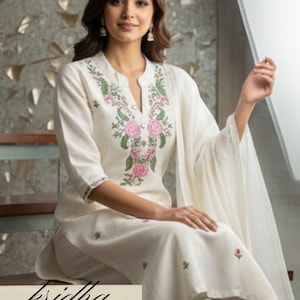 K&ouml;nnte beinhalten: Cremefarbenes besticktes Kurta-Set. Das Oberteil hat einen V-Ausschnitt mit rosa und gr&uuml;ner Blumenstickerei. Das Set enth&auml;lt eine passende Hose und ein Dupatta. Der Markenname "kridha FASHION" ist sichtbar.
