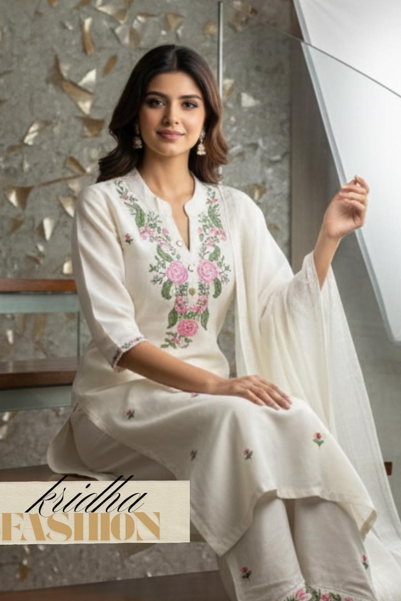 K&ouml;nnte beinhalten: Cremefarbenes besticktes Kurta-Set. Das Oberteil hat einen V-Ausschnitt mit rosa und gr&uuml;ner Blumenstickerei. Das Set enth&auml;lt eine passende Hose und ein Dupatta. Der Markenname "kridha FASHION" ist sichtbar.