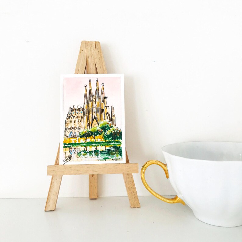 Puede incluir: Una acuarela de la Sagrada Familia en Barcelona, Espa&ntilde;a, expuesta en un peque&ntilde;o caballete de madera. La obra presenta la intrincada arquitectura de la catedral, con un reflejo en el agua. Una taza de t&eacute; blanca con asa dorada en primer plano.