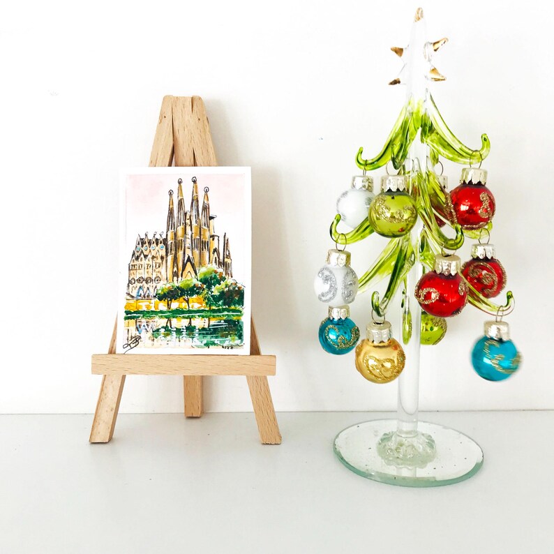 Puede incluir: Una acuarela de una catedral sobre un peque&ntilde;o caballete de madera junto a un &aacute;rbol de Navidad de cristal decorado con adornos coloridos. El &aacute;rbol tiene una estrella blanca en la parte superior y una base de cristal transparente.