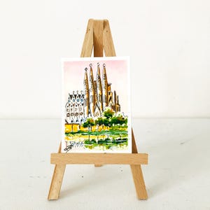 Puede incluir: Una acuarela de la Sagrada Familia en Barcelona, Espa&ntilde;a, expuesta en un peque&ntilde;o caballete de madera. La obra presenta las agujas y la intrincada fachada de la catedral, con &aacute;rboles verdes y un reflejo amarillo y verde en primer plano.