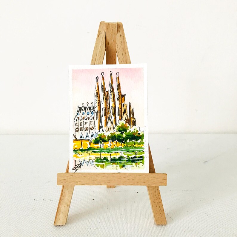 Puede incluir: Una acuarela de la Sagrada Familia en Barcelona, Espa&ntilde;a, expuesta en un peque&ntilde;o caballete de madera. La obra presenta las agujas y la intrincada fachada de la catedral, con &aacute;rboles verdes y un reflejo amarillo y verde en primer plano.