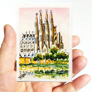 Puede incluir: Una pintura de acuarela en miniatura de la Sagrada Familia en Barcelona. La obra de arte representa las agujas de la catedral, las fachadas detalladas y la vegetaci&oacute;n circundante, con un reflejo en el agua. La pintura se sostiene en una mano.