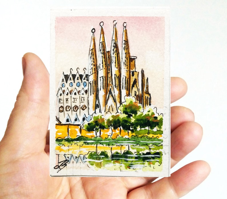 Puede incluir: Una pintura de acuarela en miniatura de la Sagrada Familia en Barcelona. La obra de arte representa las agujas de la catedral, las fachadas detalladas y la vegetaci&oacute;n circundante, con un reflejo en el agua. La pintura se sostiene en una mano.