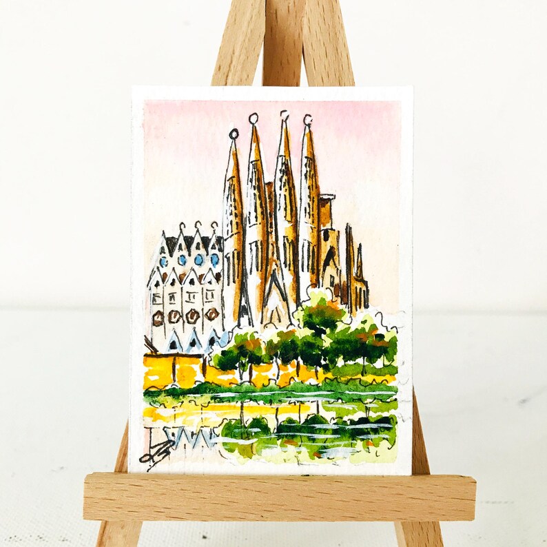 Puede incluir: Pintura a la acuarela de la Sagrada Familia en Barcelona, Espa&ntilde;a. La obra representa las intrincadas agujas y fachada de la catedral, con un reflejo en el agua debajo. El cielo es de un rosa suave y los &aacute;rboles son verdes.