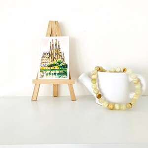 Puede incluir: Una acuarela de una catedral sobre un peque&ntilde;o caballete de madera, junto a una regadera de cer&aacute;mica blanca que sostiene una pulsera de cuentas. La pulsera presenta cuentas amarillas claras y marrones.