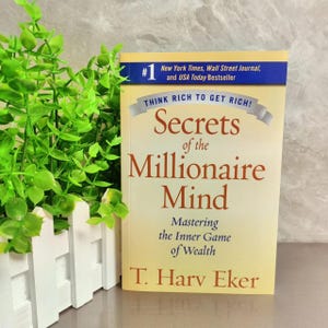 May include: A hardback book titled "Secrets of the Millionaire Mind" by T. Harv Eker. The book has a yellow cover with red and brown text. The top of the book has a blue banner that reads "New York Times, Wall Street Journal, and USA Today Bestseller."
