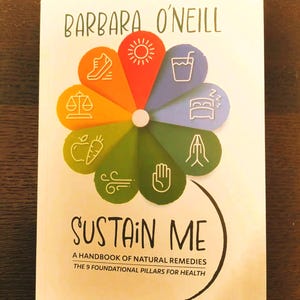 Puede incluir: Un libro titulado "SUSTAIN ME" de Barbara O'Neill. La portada presenta un diseño floral colorido con iconos que representan remedios naturales. El texto dice "A Handbook of Natural Remedies, The 9 Foundational Pillars for Health."