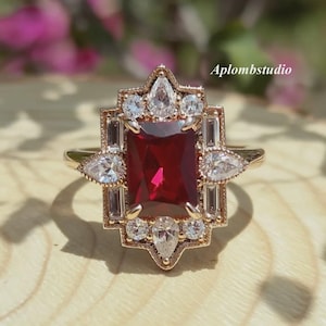 May include: A gold-toned ring featuring a large, rectangular, deep red gemstone at its center, surrounded by smaller, clear gemstones and geometric accents. The ring has a vintage, Art Deco style, with the text "Aplombstudio" visible.