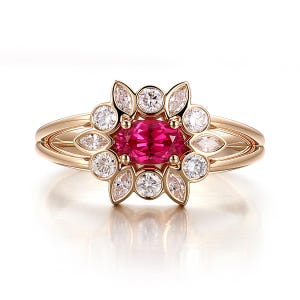 Może przedstawiać: Złoty pierścionek z centralnym owalnym rubinem, otoczonym kwiatowym układem okrągłych i markizowych diamentów. Pierścionek ma podzielony trzon. Kamienie szlachetne są osadzone w polerowanym złocie.