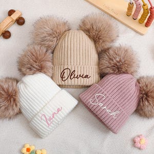 Op de afbeelding: Drie geribbelde gebreide mutsen met elk twee grote pompons van imitatiebont. De beige muts heeft de naam Olivia in bruin, de witte muts heeft de naam Luna in roze en de oudroze muts heeft de naam Sarah in wit borduursel.