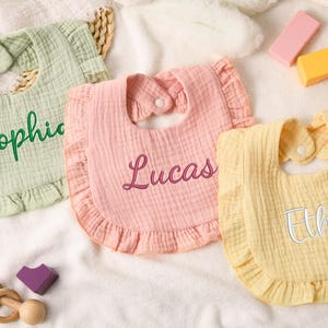 Puede incluir: Tres baberos para bebés en colores pastel: verde claro, rosa y amarillo. Cada babero tiene un borde con volantes y un cierre de botón a presión en el cuello. Los nombres "Sophie", "Lucas" y "Ethan" están bordados en los baberos.