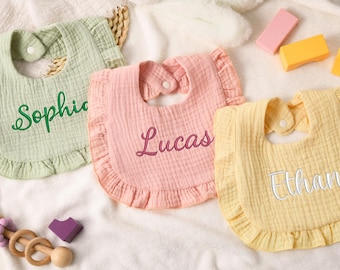 May include: Three baby bibs in pastel colors: light green, pink, and yellow. Each bib has a ruffled edge and a snap closure at the neck. The names "Sophie", "Lucas", and "Ethan" are embroidered on the bibs.