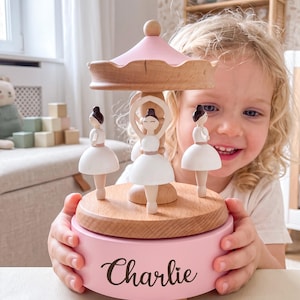 Op de afbeelding: Een houten muziekcarrousel met drie witte ballerina-figuurtjes met donkere knotjes. De basis is lichtroze geschilderd en toont de naam Charlie in zwarte schrijfletters. De bovenkant is ook lichtroze en rust op een houten middenstijl.