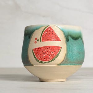 May include: A ceramic bowl with a watermelon design. The bowl features a cream-colored base and a turquoise and green glazed top. Two watermelon slices are printed on the side, with red fruit and green rind.