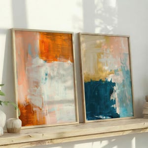 May include: Two framed abstract art prints. The artwork features a blend of orange, white, and blue hues in one, and blue, gold, and pink in the other. The frames are light-colored wood, and the art is displayed on a wooden shelf.