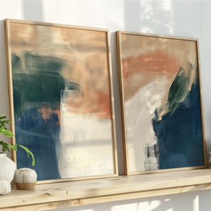 May include: Two framed abstract art prints. The artwork features brushstrokes of navy blue, teal, peach, and white. The frames are a light wood colour. The prints are displayed on a wooden shelf with plants and books.