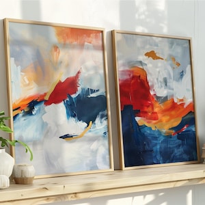 May include: Two framed abstract art prints with a wooden frame. The artwork features bold brushstrokes in shades of red, orange, blue, and white. The paintings are displayed on a wooden shelf with decorative items and plants.