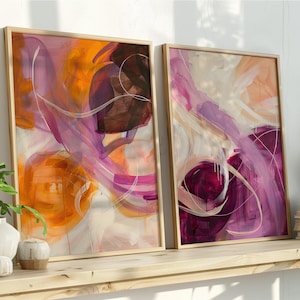 May include: Two framed abstract art prints. The artwork features flowing brushstrokes in shades of orange, purple, and white. The frames are a light wood color, and the art is displayed on a wooden shelf with decorative items.