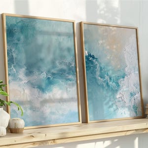 May include: Two framed abstract art prints with ocean-inspired designs in shades of blue, white, and beige. The artwork is displayed on a wooden shelf with decorative plants and books. The frames are made of light-colored wood.