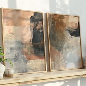May include: Two framed abstract art prints with neutral colors. The artwork features brushstrokes of beige, black, and gray. The frames are a light wood color. The prints are displayed on a wooden shelf with books and plants.