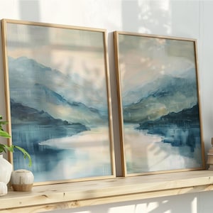 Könnte beinhalten: Zwei gerahmte Landschaftsbilder mit einer ruhigen Berg- und Seeszene in Blau-, Grau- und Beigetönen. Das Kunstwerk wird auf einem Holzregal mit dekorativen Gegenständen und Pflanzen ausgestellt. Die Rahmen sind hellholzfarben.
