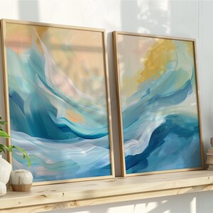 May include: Two framed abstract art prints with a watercolor effect. The artwork features flowing blue, yellow, and peach tones. The frames are light wood. The prints are displayed on a wooden shelf with plants and books.