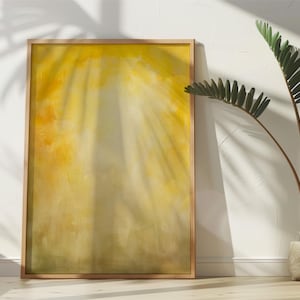 Pode incluir: Uma obra de arte abstrata emoldurada com uma paleta de cores amarelas e douradas, encostada a uma parede branca. A obra de arte tem uma apar&ecirc;ncia suave e desfocada, com efeitos de luz e sombra. Uma planta em vaso com folhas verdes est&aacute; &agrave; direita.
