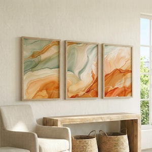 May include: A set of three rectangular wall art prints featuring abstract fluid marble patterns in shades of sage green, cream, and burnt orange. Each print is encased in a light-toned wooden frame, displaying flowing, organic color gradients.