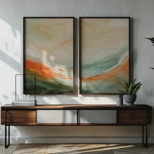 May include: Two framed abstract artworks with orange, green, and beige hues are displayed above a dark wood cabinet. A potted plant and decorative items sit on the cabinet. A large potted plant with green leaves is on the right.