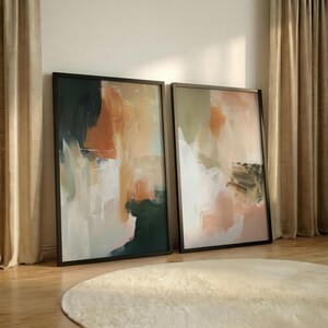 Könnte beinhalten: Zwei gerahmte abstrakte Kunstwerke. Das Kunstwerk zeigt eine Mischung aus Farben, darunter Orange, Weiß, Grün und Schwarz. Die Rahmen sind schwarz und die Kunst wird vor einer neutralen Wand ausgestellt. Die Stücke lehnen an der Wand.