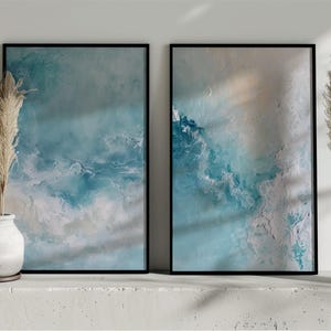 May include: Two framed abstract art prints with ocean-inspired designs in shades of blue, white, and beige. The artwork is displayed on a white surface, complemented by decorative pampas grass in white vases. The frames are black.