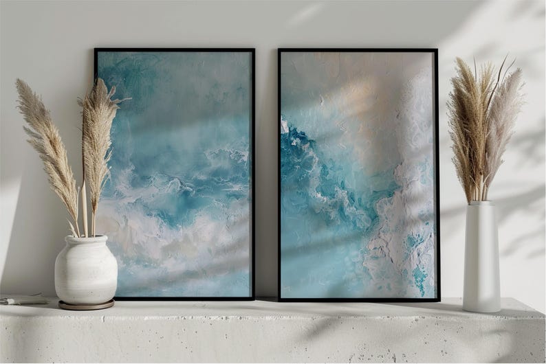 May include: Two framed abstract art prints with ocean-inspired designs in shades of blue, white, and beige. The artwork is displayed on a white surface, complemented by decorative pampas grass in white vases. The frames are black.