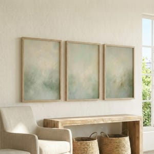 May include: Three framed abstract art prints with a soft, muted color palette of greens, creams, and yellows. The artwork is displayed above a wooden console table, with a neutral-toned armchair and woven baskets in the foreground.