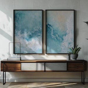May include: Two framed abstract art prints with blue and white tones hang above a wooden console table. A potted plant and decorative items sit on the table. A large potted plant with green leaves is to the right of the table.